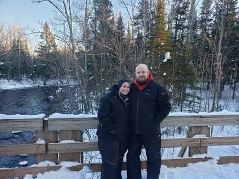 Kim and Alex are standing on a bridge in the wintertime in snowmobile gear