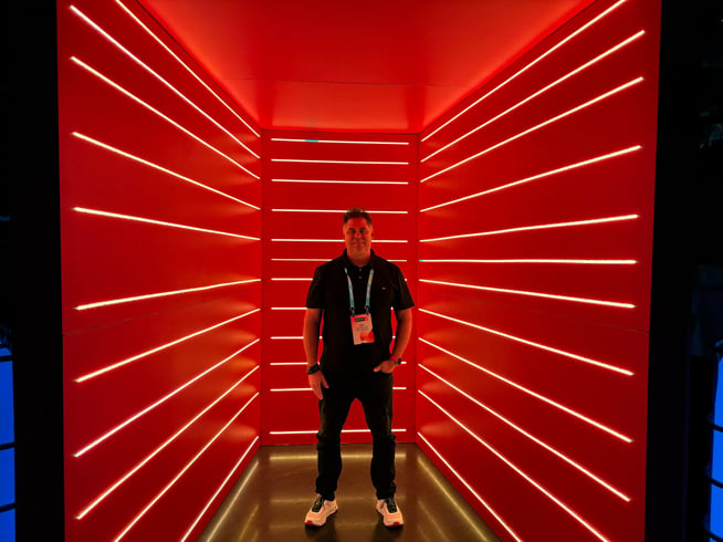 Man in dark shirt, jeans and white sneakers stands with left hand in pocket inside 3 red walls with horizontal LED lines going across.