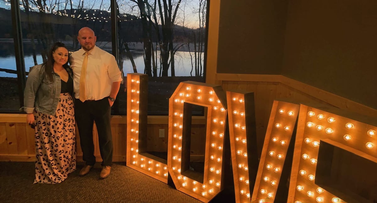 Kim and Alex at a wedding, standing in front of large, lit up letters that spell love