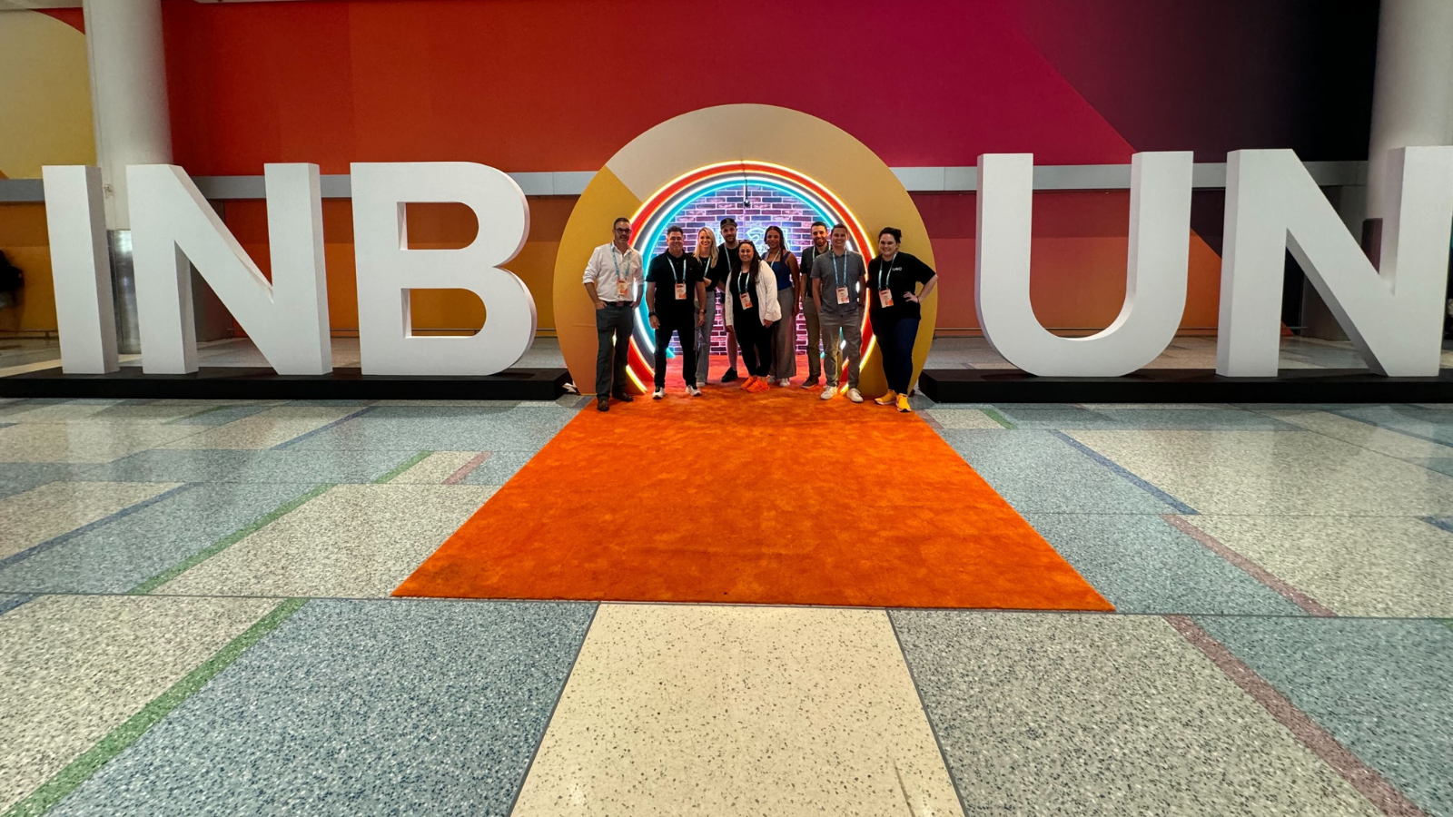 Group of nine people stand on an orange carpet in front open O in a sign that spells out INBOUND.